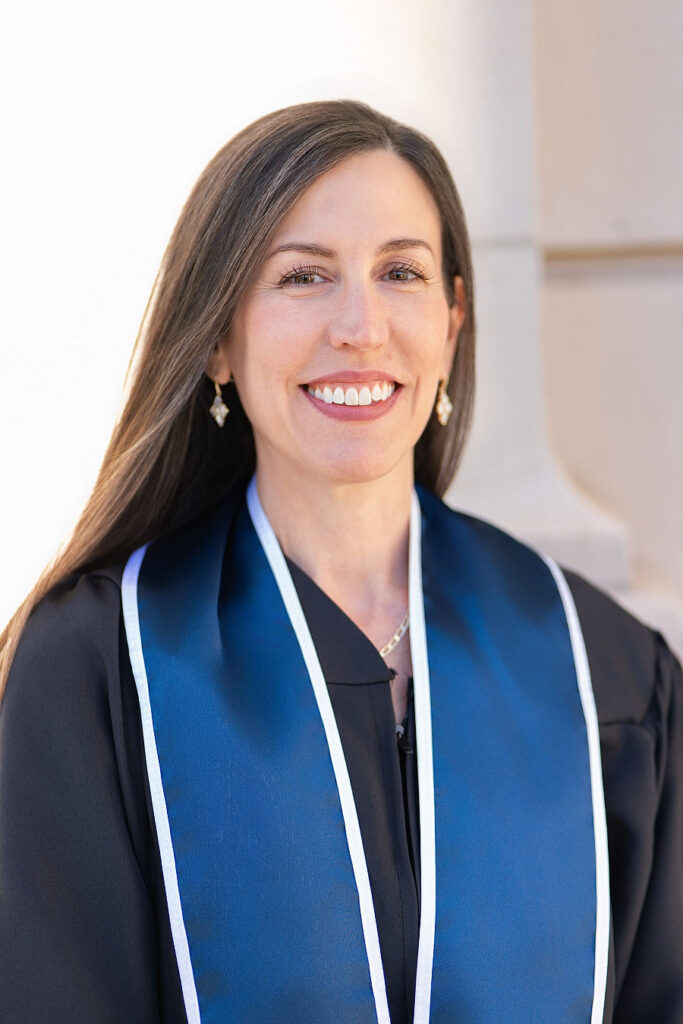ACC alumna Claire Hobbs smiling in a black graduation robe with a dark blue satin stole trimmed in white.