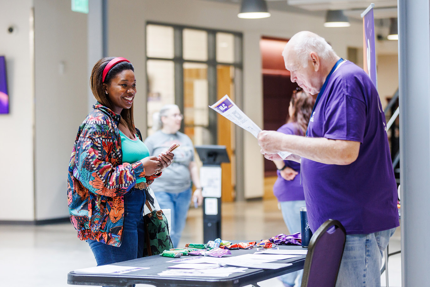 Welcome Riverbats - Fall 26 Here is an alt text description for the image: “A smiling Black woman in a colorful jacket and a red headband is engaged in conversation with an older gentleman wearing a purple shirt at a table covered with promotional materials. Several other people are visible in the background, suggesting a registration or information booth setting.”