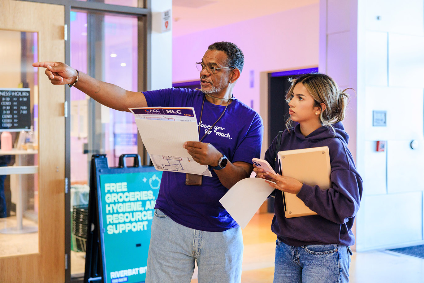Welcome Riverbats - Fall 26 Here is an alt text description for the image: “A Black man in a purple shirt and glasses is pointing to a document while speaking to a young woman in a purple shirt. A sign behind them reads “FREE GROCERIES, HYGIENE, AND RESOURCE SUPPORT” with the Riverbat logo. The setting appears to be an indoor space with a large window and a muted purple wall.”