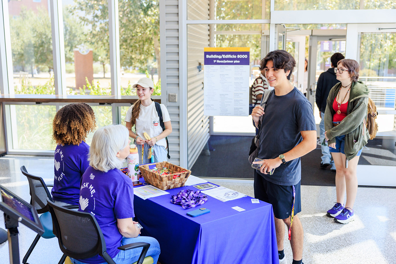 Welcome Riverbats - Fall 26 Here’s an alt text description for the image: “A young man in a blue t-shirt and shorts is speaking with a woman at a purple table in a brightly lit hallway. Two other students stand nearby, observing the interaction. A sign displaying building information is visible in the background.”