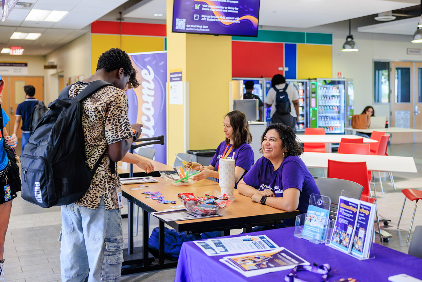 Welcome Riverbats - Fall 26 Here’s an alt text description for the image: “A young Black man stands at a table covered with purple promotional materials, speaking with a smiling Hispanic woman wearing a purple shirt. Several other students are visible in the background. The table is set in a brightly colored cafeteria with a large digital screen displaying information about the university.”