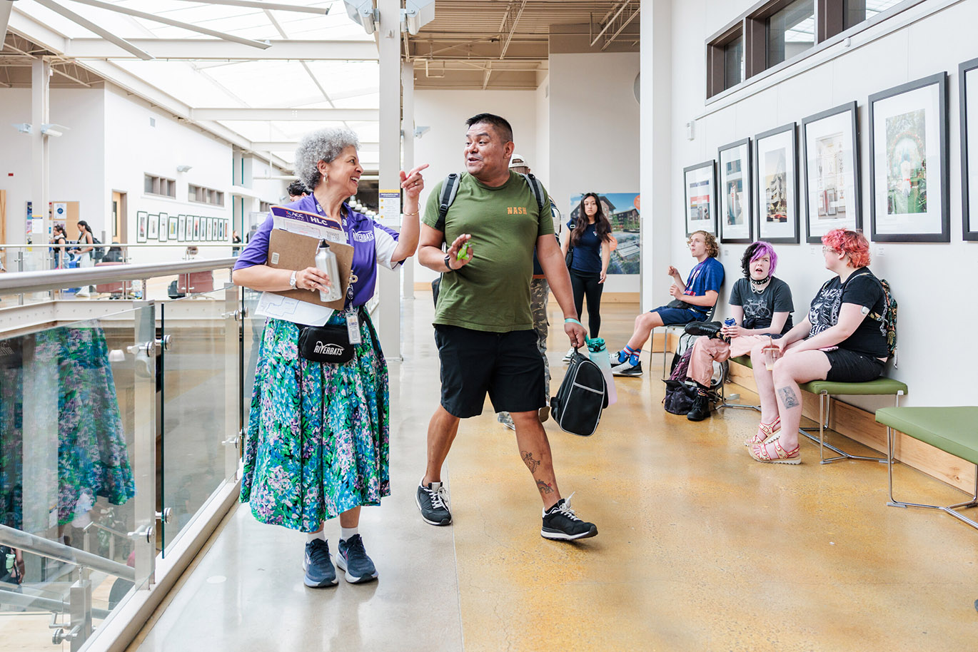 Welcome Riverbats - Fall 26 Here’s an alt text description for the image: “A diverse group of people, including an elderly woman in a patterned skirt and a man in a camouflage shirt, are walking through a brightly lit hallway in a museum. Several other individuals are seated on benches, observing the artwork displayed on the walls. The artwork consists of framed photographs of landscapes.”