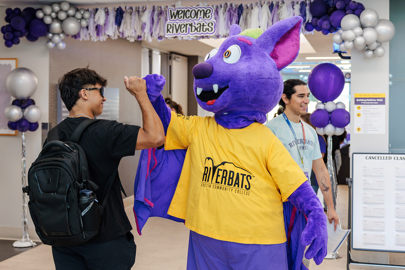 Welcome Riverbats - Fall 26 Here's an alt text description for the image: “A purple River Bats mascot, a cartoon bat with a smiling face and large eyes, extends a paw in a high-five to a young man wearing a yellow River Bats t-shirt. The man is standing next to a young man in a River Bats t-shirt, and a backdrop of purple balloons and streamers is visible.”