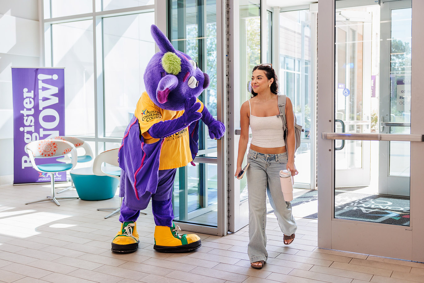 Welcome Riverbats - Fall 26 Here’s an alt text description for the image: “An Allegonaut mascot, a purple and green rabbit, stands next to a glass door entrance. A young woman in a white tank top and light wash jeans walks past, carrying a backpack and holding a phone in her hand. The background is a modern, brightly lit university hallway with a “Register NOW!” banner.”