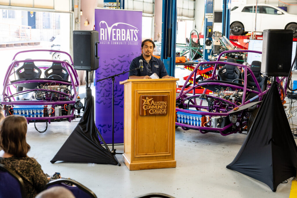A man stands at a podium with the logo of Austin Community College, speaking to an audience in a garage-like setting. Behind him are two purple off-road vehicles, and there are black drapes on either side of the podium. The environment suggests a presentation or event related to automotive technology or education.